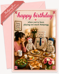 A birthday card and matching pink envelope shown on a white background. The card features a photo of a woman dining with three mahjong-themed robot figures and gift boxes, with the text 'happy birthday when you've been playing too much Mahjongg'.