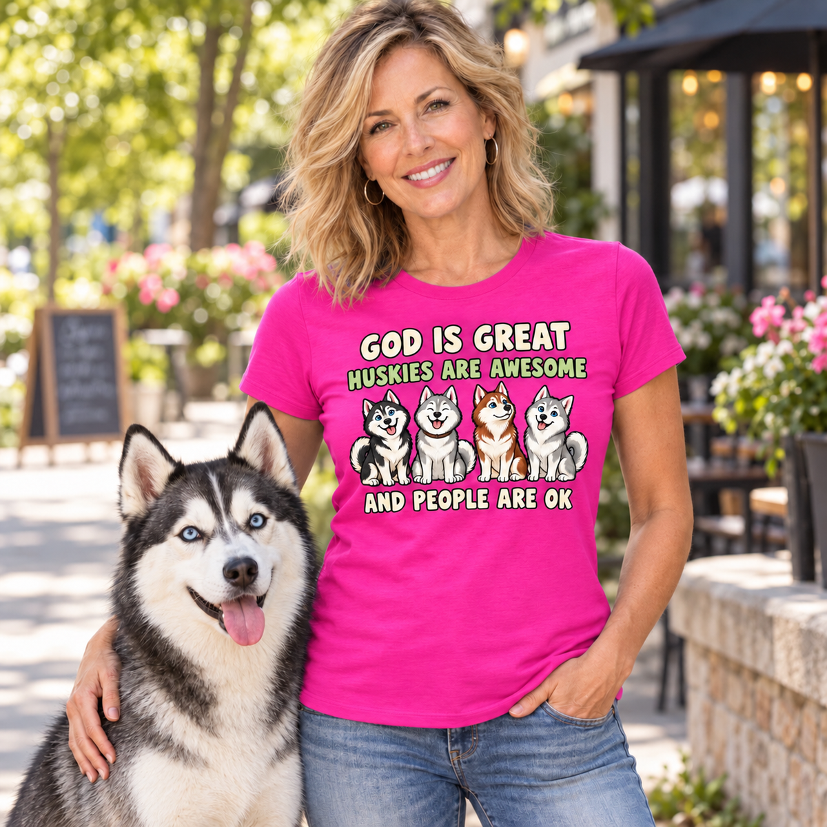 A woman wearing a bright pink graphic t-shirt with a husky-themed slogan and illustrations, standing outdoors next to a husky dog.