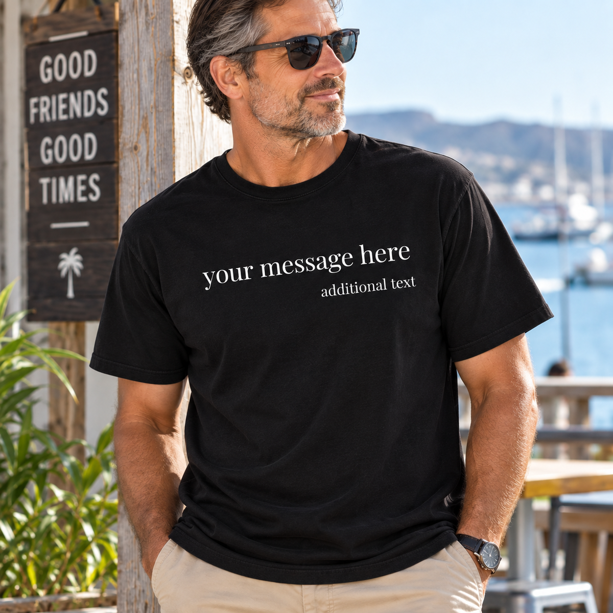 A man wearing a black customizable graphic t-shirt with white placeholder text, standing outdoors near a wooden sign and a sunny harbor background.