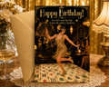 A personalized 1920s flapper-themed birthday card with an envelope, displayed on a wooden table with a champagne glass, lace doily, and lamps in the background.
