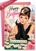 A pink and gold Happy Birthday greeting card featuring an illustrated portrait of Audrey Hepburn in a black dress and gloves, surrounded by mahjong tiles, flowers, and a teacup, with a pink envelope behind it.