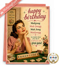 A square birthday card featuring a retro illustration of a woman playing mahjong, with pink borders and the text 'happy birthday Mahjong Mah Jongg' on a cream background. A pink envelope is partially visible behind the card, and a 'MahjongFun' ribbon and an 'Seen on NBC' badge appear around the edges.