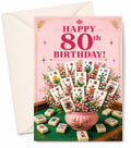 A pink 80th birthday greeting card with a cream envelope behind it. The card features the text 'HAPPY 80th BIRTHDAY!' above an illustration of mahjong tiles arranged like flowers in a pink pot, with additional mahjong tiles scattered around on a green table.