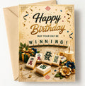 A gold and beige 'Happy Birthday' greeting card with a kraft envelope behind it. The card features a mahjong-themed design with wooden tile graphics spelling 'WINNING!', decorative flowers, a candle, and a gift box.