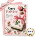 A pink and white Happy Birthday greeting card featuring a mahjong-themed design with a gift box wrapped in a pink ribbon, scattered mahjong tiles, and pink roses. A pink ribbon banner reads 'MahjongFun', and a gold 'As Seen On' badge is in the bottom right corner.