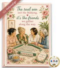 A floral greeting card or art print featuring four women gathered around a mahjong table, with decorative pink flowers and the text, 'The real win isn't the Mahjong, it's the friends we gather along the way.' A pink ribbon banner reads 'Mahjong fun!' and a round seal says 'As seen on The NBC with ABC.