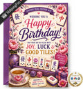 A square birthday card design with a Chinese mahjong theme, featuring floral artwork, playing card tiles, a teacup with pink flowers, gift boxes, and celebratory text including 'Happy Birthday!' and 'May your day be filled with joy, luck & good tiles!'. A black diagonal banner reads 'MahjongFun' and a circular badge says 'It's seen on TV'.