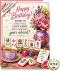 A colorful Happy Birthday greeting card or print featuring mahjong-themed illustrations, including floral pink and purple peonies in a vase, several decorated mahjong tiles, dice, a teacup, and decorative text wishing good tiles and lucky hands.