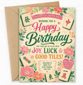 A floral mahjong-themed birthday card with a brown kraft envelope behind it, photographed on a light neutral background. The card features pink and green text reading 'Happy Birthday' with decorative flowers and several illustrated mahjong tiles.